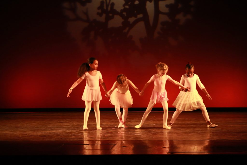 Eine Gruppe von jungen Balletttänzerinnen in weißen und rosa Outfits führt eine synchronisierte Bewegung im Tanzstudio München aus.