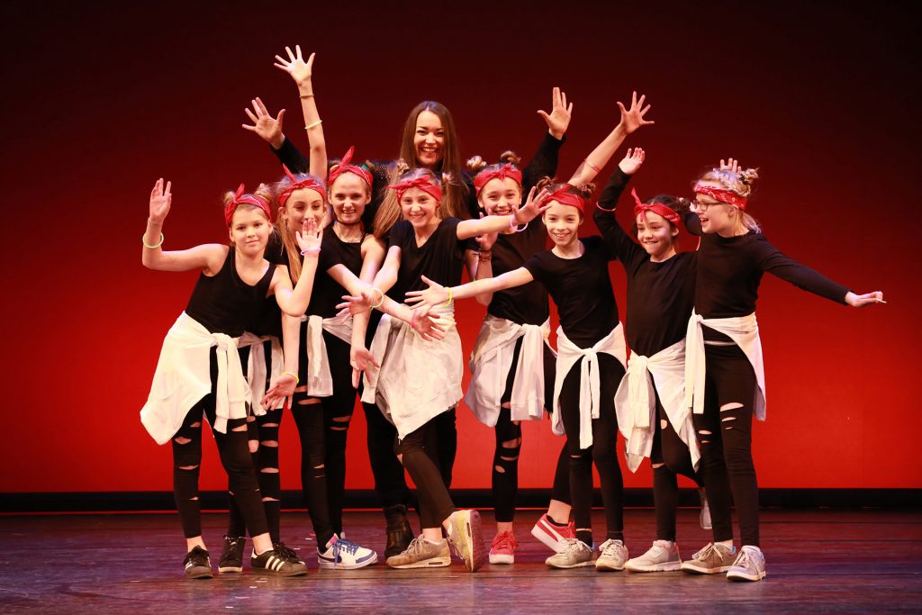 Eine Gruppe junger Hip-Hop Tänzerinnen in schwarz-roten Outfits mit weißen Sweatshirts posiert fröhlich auf der Bühne im Tanzstudio München.
