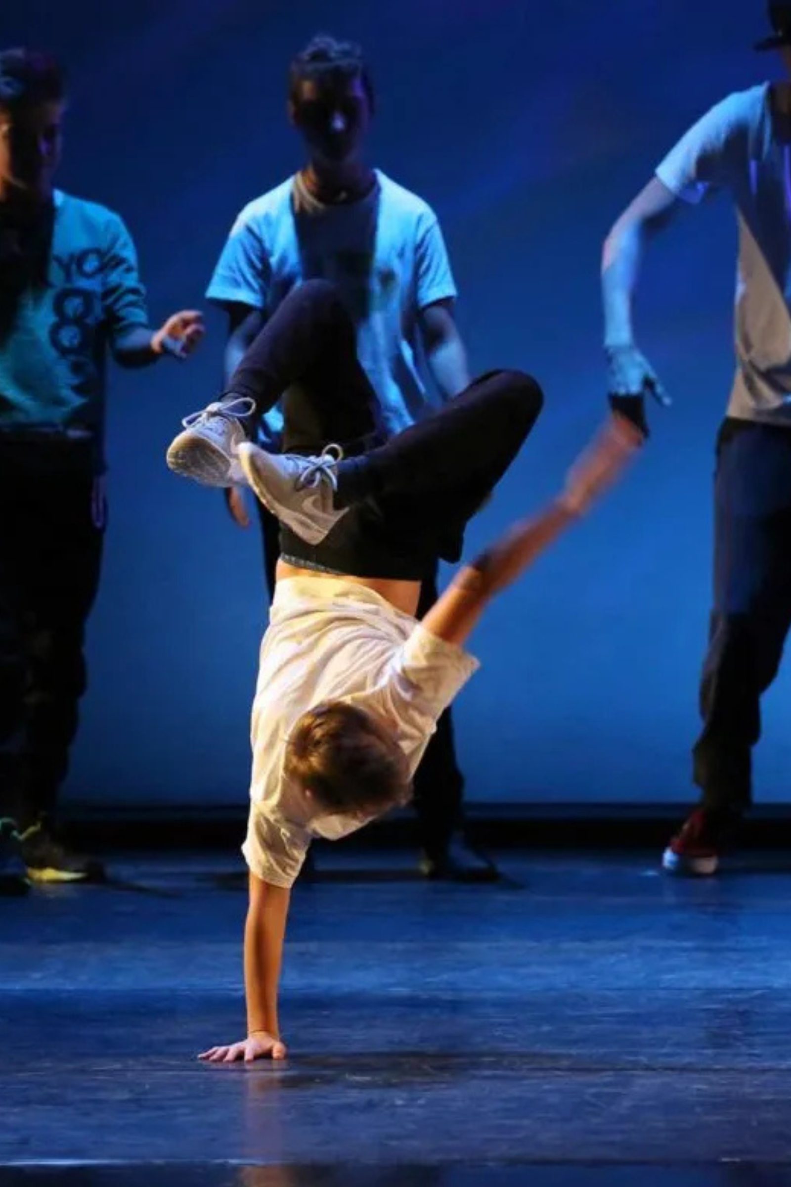 Breakdance Schüler bei KayJay’s Dance Studio in München zeigt einen einarmigen Handstand auf der Bühne, während andere Tänzer im Hintergrund zusehen.