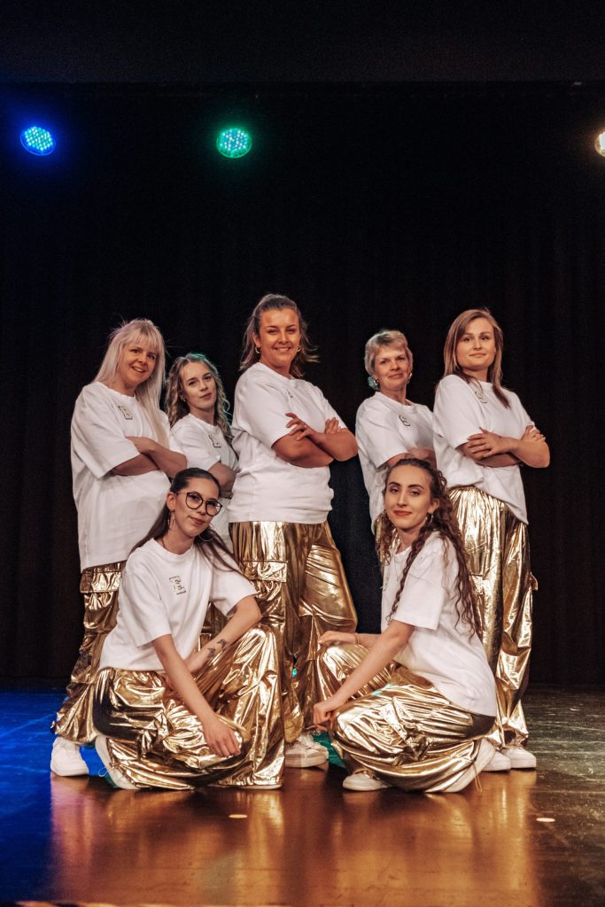 Gruppe Erwachsener in weißen Shirts und goldenen Hosen auf der Bühne