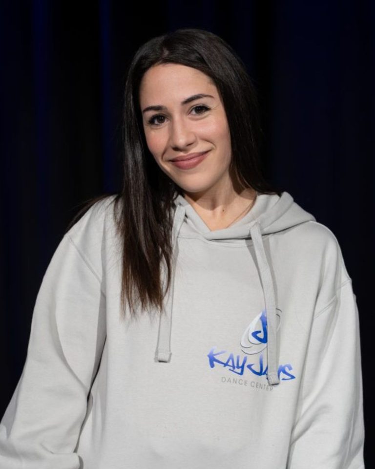 Rania, Trainerin bei KayJays, im hellgrauen KayJays-Hoodie vor blauem Bühnenvorhang